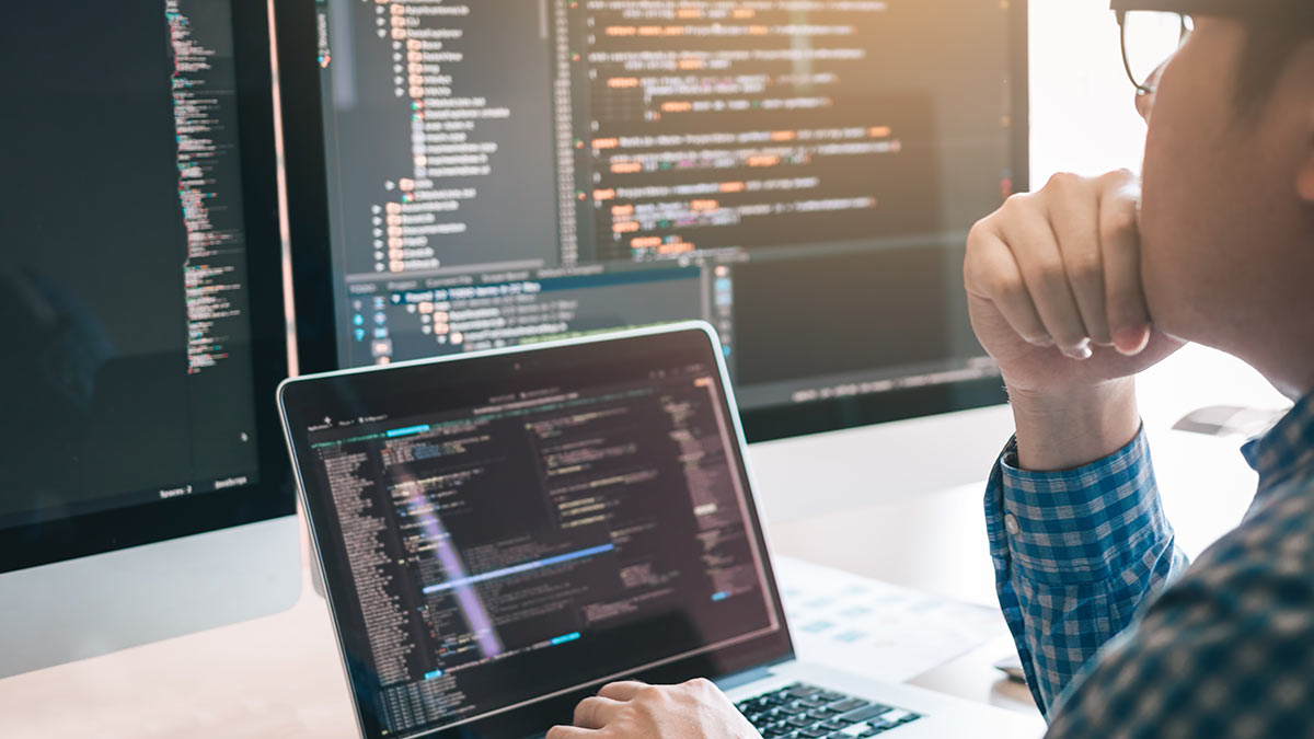 A person with glasses looking at a laptop and another screen with code on them.