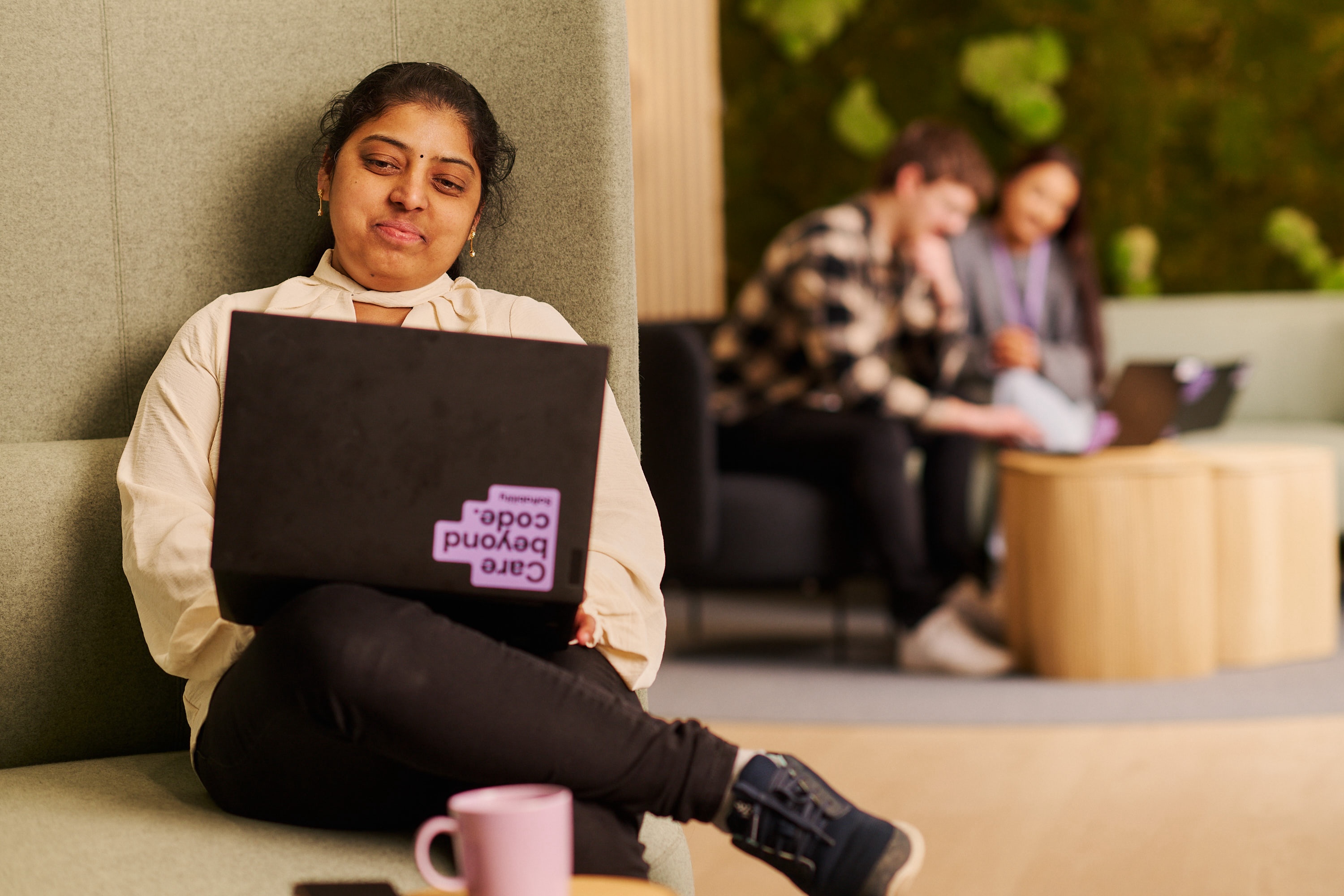 A woman sitting in a lounge chair with a laptop facing the camera. Two people in the background working.
